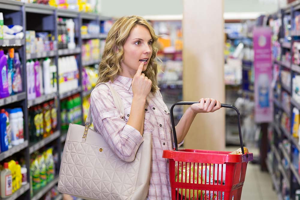 Жінка із сумкою для покупок задумливо дивиться на полицю у супермаркеті.